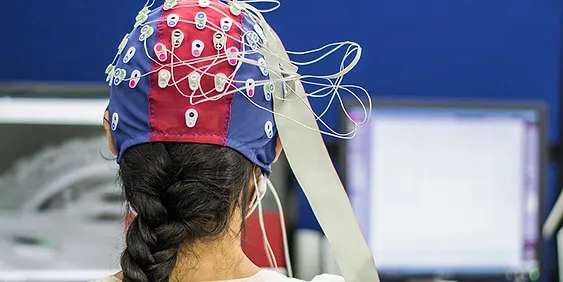 A person with long brown hair in a braid sits facing away from the camera, wearing a colorful EEG cap with numerous electrodes and wires attached. Two blurry computer screens are in the background, indicating a possible neuroscience or medical research setting.