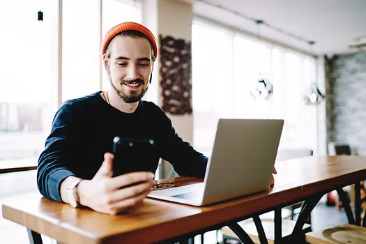 A smiling man with a beard, wearing an orange beanie and black sweater, sits at a large wooden table with a laptop in front of him. He is holding and looking at his phone. The background features large windows letting in natural light.