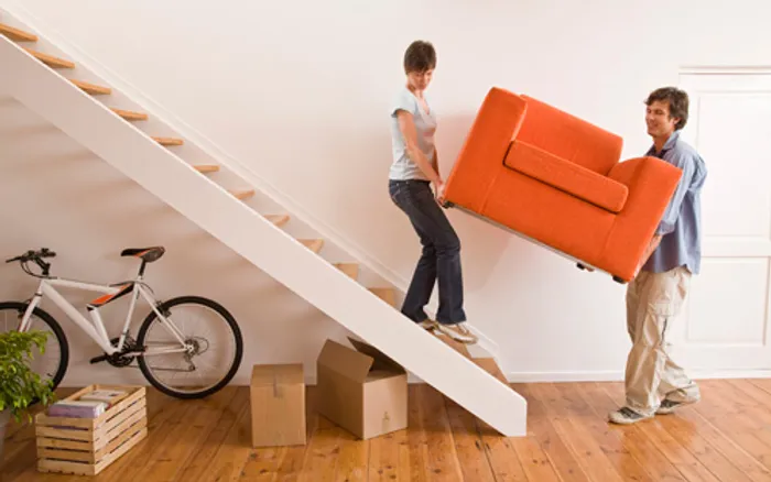 A man and a woman are carrying an orange armchair up a wooden staircase in a bright, minimally decorated room. Two cardboard boxes, a white bicycle, and a small stack of books are on the floor near the staircase.