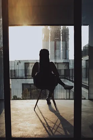 Silhouette of a person with braided hair sitting on a chair, facing a large window. Sunlight shines through the window, casting shadows on the floor. The backdrop features an industrial structure, with some buildings and an urban skyline visible in the distance.