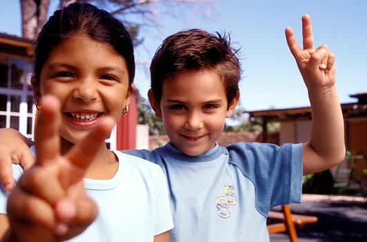 Smiling girl and boy standing close together outside, both holding up peace signs with their fingers. The sky is clear, and a building is visible in the background. The girl has dark hair and the boy has light brown hair, both wearing light-colored shirts.