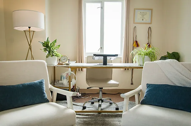 A cozy home office features a desk with a computer monitor, surrounded by green plants and a certificate on the wall. Two upholstered chairs with blue cushions face the desk, and a tall lamp stands in the corner. A window lets in natural light.