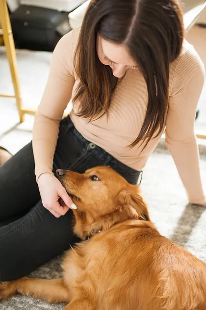 A woman with long brown hair, wearing a beige long-sleeve top and black pants, is sitting on the floor and gently petting a golden retriever. The dog is lying down and looking up at the woman affectionately.