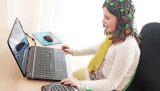 A young girl sits at a desk focused on a laptop screen. She wears a green EEG cap with electrodes, and a yellow scarf over a white sweater. Her right hand is on a circular device next to the laptop. A mouse pad and wireless mouse are visible on the desk.
