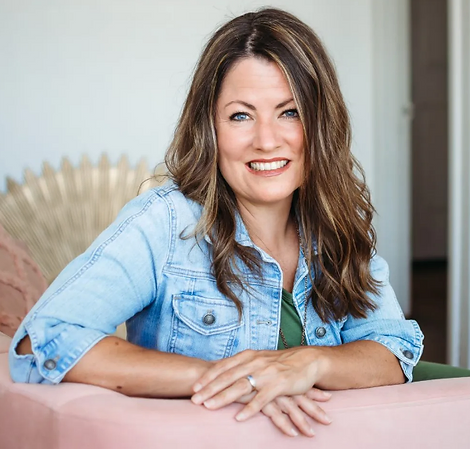 A woman with long brown hair, wearing a denim jacket over a green top, smiling and leaning forward slightly while resting her arms on the back of a pink couch. She is indoors, and a sunburst-style decor is visible in the background.