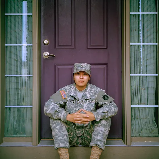 A person in military uniform is sitting on the steps in front of a brown door with their hands resting together in their lap. The door is flanked by windows, and the individual has a calm expression, wearing a cap and camouflage attire.
