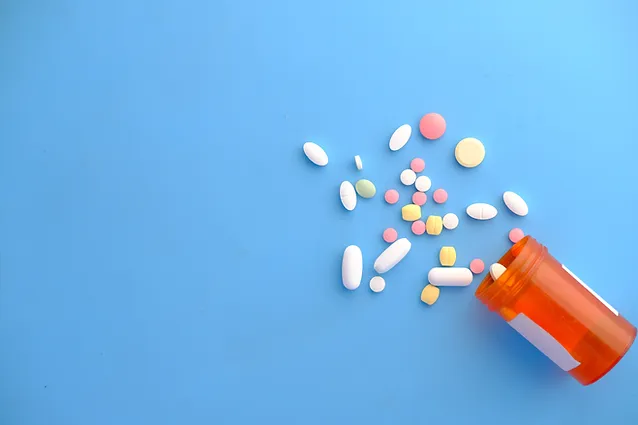 A variety of pills in different shapes and colors spill out of an orange prescription bottle on a light blue background. The pills include white, pink, yellow, and multicolored tablets and capsules.