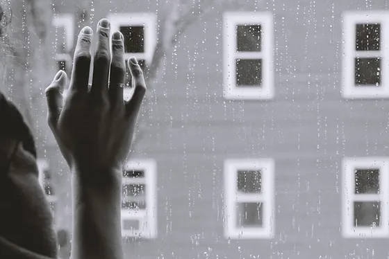 A hand gently touches a rain-streaked window, with several white-framed windows of an adjacent building visible outside. The black-and-white image evokes a sense of contemplation or longing as raindrops blur the structures in the background.