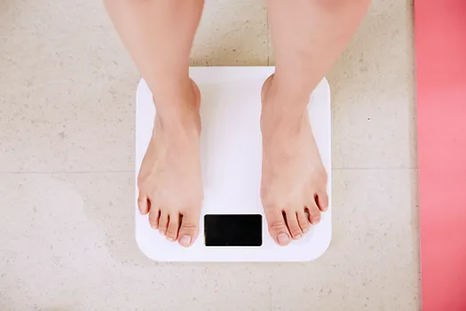 A person standing on a white digital bathroom scale with a black display screen. The scale is placed on a light-colored tiled floor, and only the person's feet and lower legs are visible. There is a glimpse of a pink mat to the right.