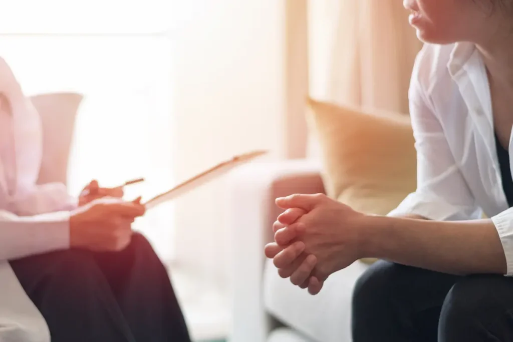 Close-up of a patient's clasped hands resting on their lap during a one-on-one therapy consultation, with a clinician holding a clipboard in the background.