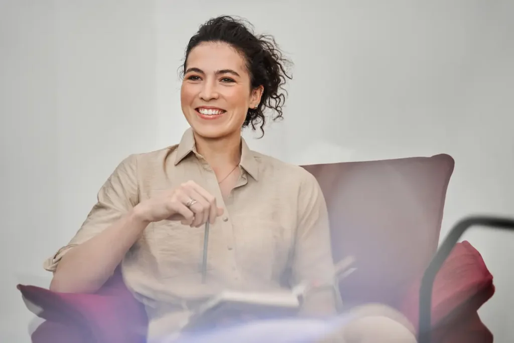Smiling female therapist with dark curly hair sitting in a comfortable chair holding a pen and notebook, representing a welcoming therapeutic environment.