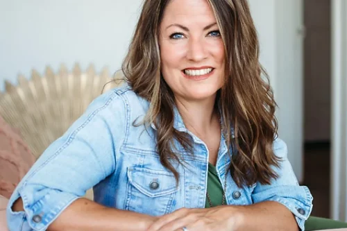 A woman with long brown hair, wearing a denim jacket over a green top, smiling and leaning forward slightly while resting her arms on the back of a pink couch. She is indoors, and a sunburst-style decor is visible in the background.